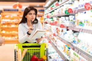 Woman thoughtfully browsing grocery store shelves with a notepad and pen in her cart representing the mental effort of managing food noise while making healthy food choices