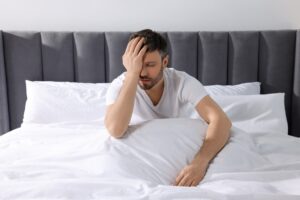 Man sitting up in bed with his hand over his face looking exhausted representing the connection between poor sleep and weight gain through disrupted appetite hormones and fatigue