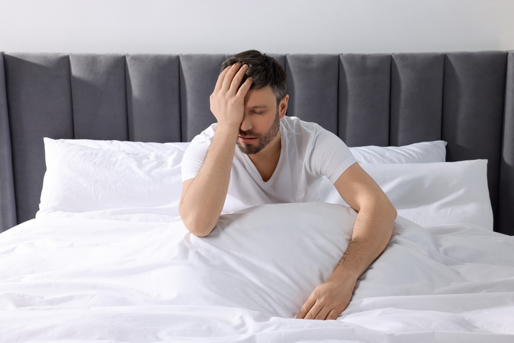Man sitting up in bed with his hand over his face looking exhausted representing the connection between poor sleep and weight gain through disrupted appetite hormones and fatigue