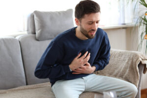 Man sitting on a couch clutching his chest with a pained expression representing concerns about testosterone and heart health and cardiovascular risk in men considering TRT