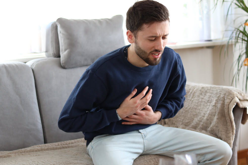 Man sitting on a couch clutching his chest with a pained expression representing concerns about testosterone and heart health and cardiovascular risk in men considering TRT