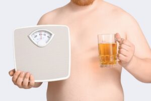 Overweight man holding a bathroom scale in one hand and a glass of beer in the other representing the conflict between alcohol and semaglutide during a medical weight loss program