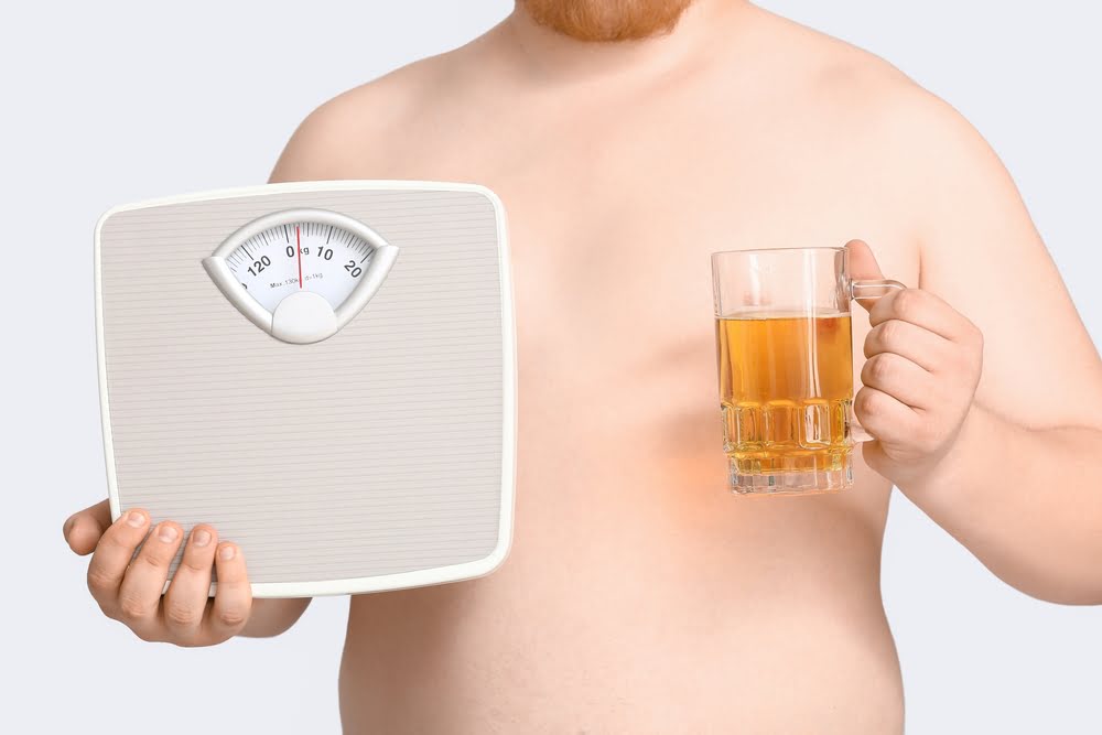 Overweight man holding a bathroom scale in one hand and a glass of beer in the other representing the conflict between alcohol and semaglutide during a medical weight loss program