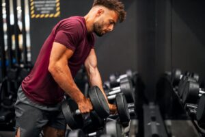 Muscular man in a red shirt lifting heavy dumbbells at the gym representing the physical results possible with TRT and muscle building through consistent resistance training