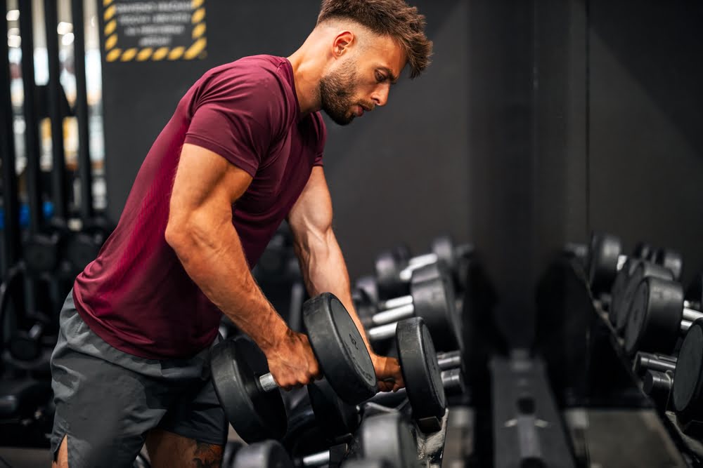 Muscular man in a red shirt lifting heavy dumbbells at the gym representing the physical results possible with TRT and muscle building through consistent resistance training