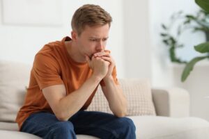 Young man in an orange shirt sitting tensely on a couch with his hands clasped to his mouth representing the anxiety and emotional changes men experience when they stop TRT