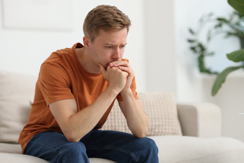 Young man in an orange shirt sitting tensely on a couch with his hands clasped to his mouth representing the anxiety and emotional changes men experience when they stop TRT