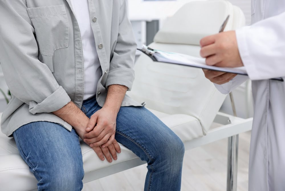 Man sitting on an exam table at a medical consultation with a provider holding a clipboard representing concerns about TRT and prostate health before starting testosterone therapy