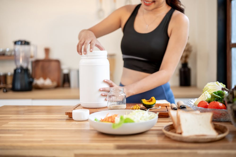 Woman in athletic wear scooping protein powder in a kitchen surrounded by fresh vegetables representing the importance of protein and weight loss strategies while on GLP-1 medications