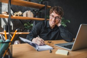Young man with glasses smiling while writing notes at a desk with a laptop representing improved testosterone and cognitive function including focus memory and mental clarity