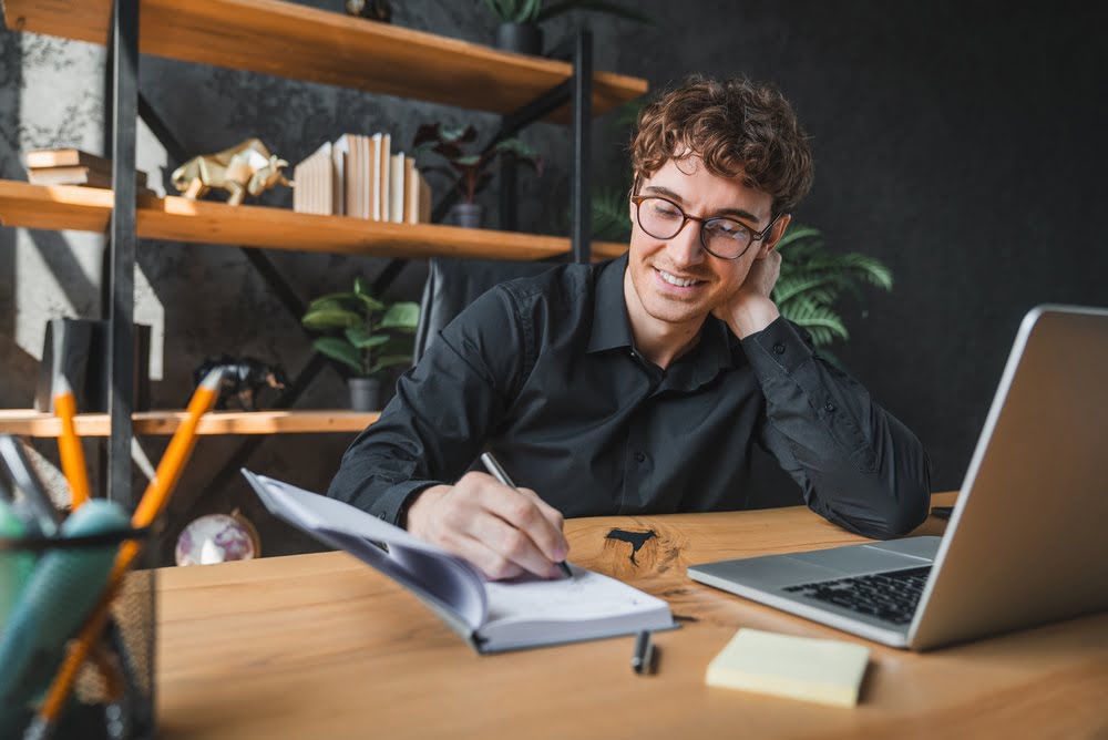Young man with glasses smiling while writing notes at a desk with a laptop representing improved testosterone and cognitive function including focus memory and mental clarity