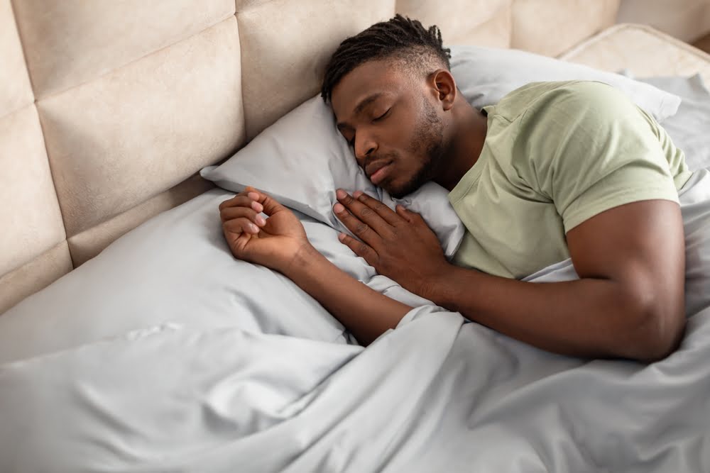 Man sleeping peacefully on his side in a bed with white and gray bedding representing the improved testosterone and sleep quality that comes with properly managed hormone levels